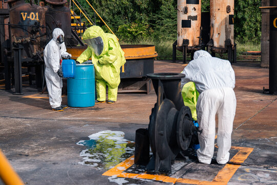 Hazmat team cleaning a chemical spill around industrial pump equipment and storage drums during an emergency response exercise at an outdoor industrial facility in protective suits