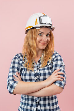 Female construction worker with blonde hair and a safety hardhat crossing arms, looking at the viewer with a confident and a little smirking expression against a plain pink background