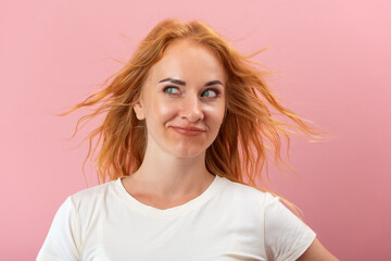 Fototapeta premium Red haired woman with blowing hair and green eyes looking mischievously to the side, expressing curiosity and a playful attitude against a pink background