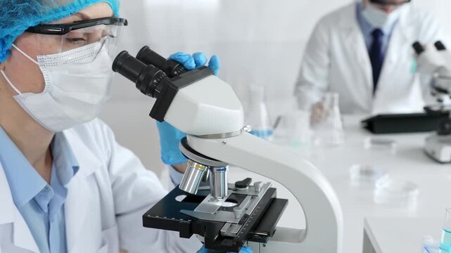 Female scientist wearing a mask and blue safety gear, using a microscope for research while a male colleague works in the background. Medicine, healthcare and science concept