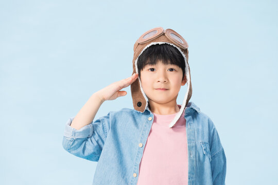 Cute young boy in aviator hat saluting on light blue background