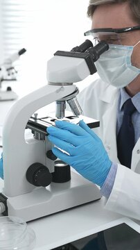 Male masked scientist wearing blue protective gloves, is using microscope doing scientific research in a laboratory with test tubes containing blue liquid. Medicine, healthcare and science concept