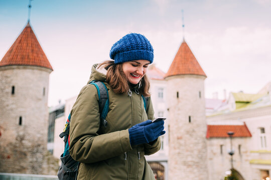 Tallinn, Estonia. Young Caucasian Woman Backpacker Tourist Enjoying Life, Smiling And Using Smartphone Near Landmark Viru Gate. Vacation In Old Town. Girl Surfing Net Internet In Phone
