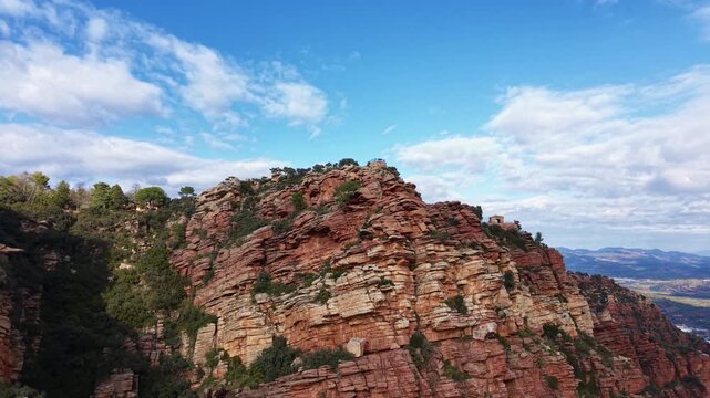 Aerial view of dramatic red sandstone cliffs at the Mirador de Garbi in the Sierra Calderona Natural Park, Valencia, Spain, with rugged rock layers, green vegetation, and a wide mountain backdrop