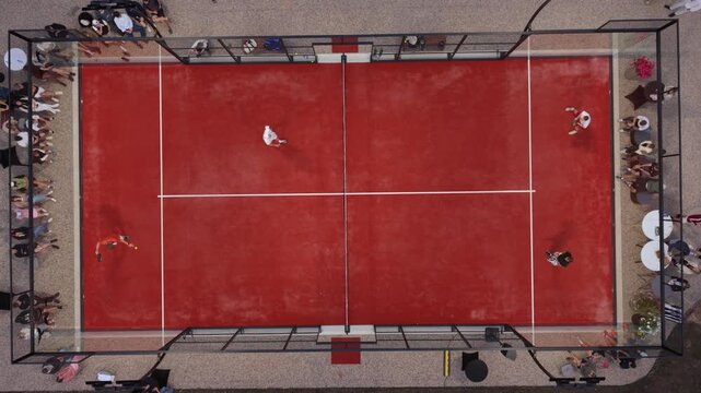 Two players engage in a competitive match on a red paddle tennis court with spectators watching from the sidelines
