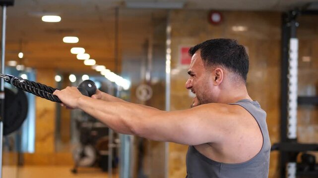 Focused athletic man performing cable face pulls exercise with intense facial expression in a modern indoor gym setting