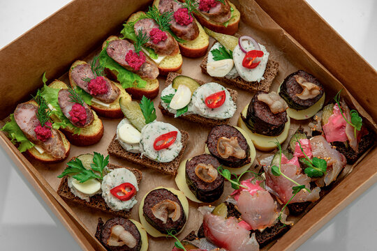Assortment of small open face sandwiches in paper catering box. Mini crostini topped with sausages, fish, pickles, and greenery are arranged inside cardboard box.