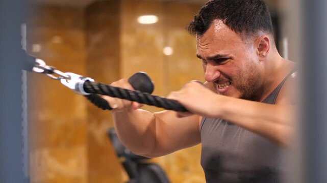 Focused athletic man training upper body with cable crossover rope extension in a modern gym, close-up shot showing sweat and intense concentration