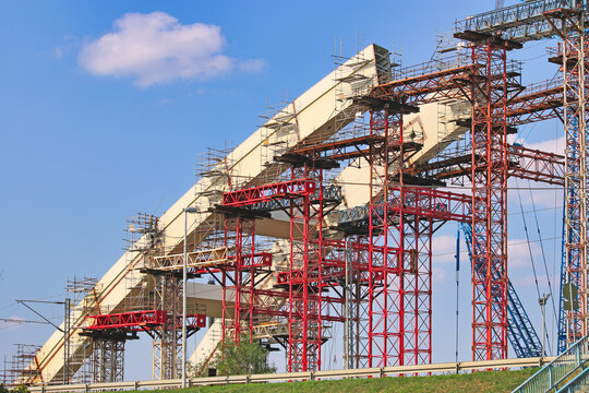 Construction site of a major steel arch bridge with complex scaffolding and temporary support towers under a blue sky