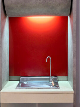Modern water drinking fountain set in a minimalist wall niche. Features a sleek chrome faucet, stainless steel basin, and a bold red background illuminated by a warm orange glow from the top light.