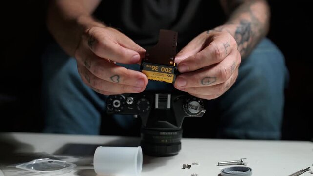 Close up of a man's tattooed hands taking a 35mm film roll out of its canister. A disassembled vintage camera is on the table