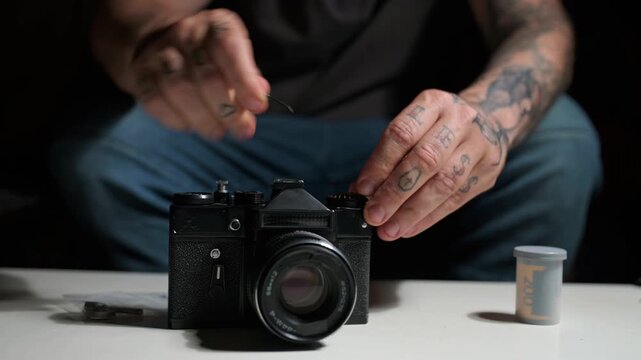 Man with tattooed arms and hands carefully repairing a vintage black analog camera on a white table. The technician uses small tools for maintenance