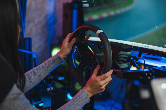 Woman playing game on car simulator while holding steering wheel in dark room with colorful neon lights, enjoying virtual drive during hobby time at home, focused on digital race track screen