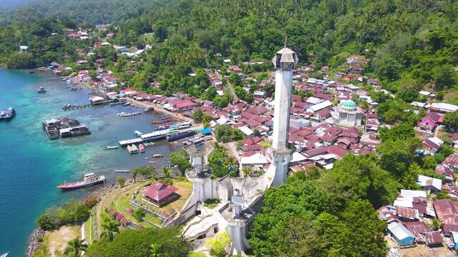 Aerial view of Batu Lubang, Lembeh Island in Bitung, Indonesia. The iconic Trikora Monument stands tall above a vibrant fishing village and boats moored in crystal-clear turquoise waters.