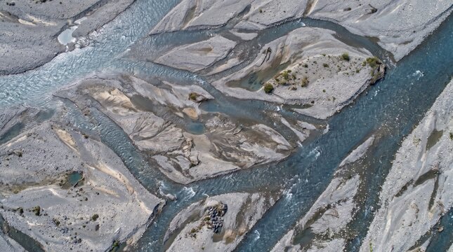 Aerial view of a braided river system with flowing blue water channels, grey gravel bars, and sediment deposits forming intricate patterns across a wide, dry riverbed landscape