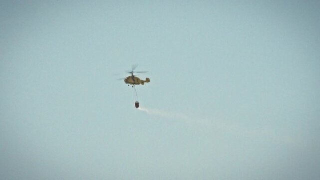 An aviation firefighting helicopter flies in a clear blue sky, its external tank full of water, conducting aerial fire suppression as part of an emergency response over an active wildfire