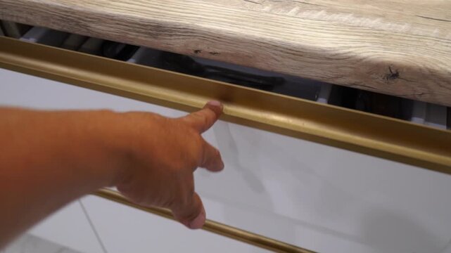 A hand reaches out to open a kitchen drawer. Inside are various utensils and cooking tools, arranged neatly. Items include forks, knives, spoons, cooking spoons, and scissors. The drawer design 