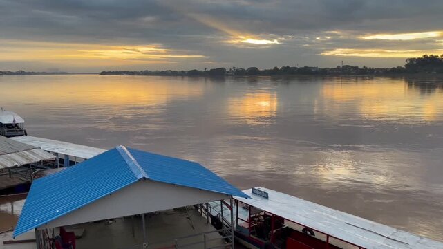 Beautiful golden sunset reflecting on calm river surface near riverside pier structure with blue roof building, showcasing peaceful scenic travel destination