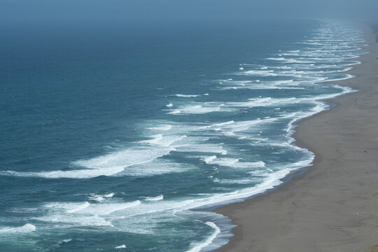 Aerial view of long sandy beach with rhythmic rolling waves and misty horizon, evoking calm and vastness. Point Reyes National Seashore, California, USA.