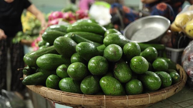 Close-up of fresh ripe green avocados displayed in a wicker basket on a stall at Xom Moi Market in Nha Trang, Vietnam. Commercial food supply and retail trade concept.