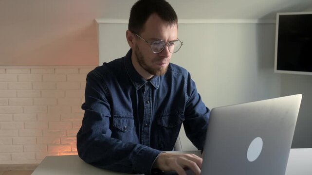 Man working on laptop in home office showing changing reactions and focused engagement over brief interaction, with man, male, portrait, glass detail, showing beard, denim, shirt, laptop context