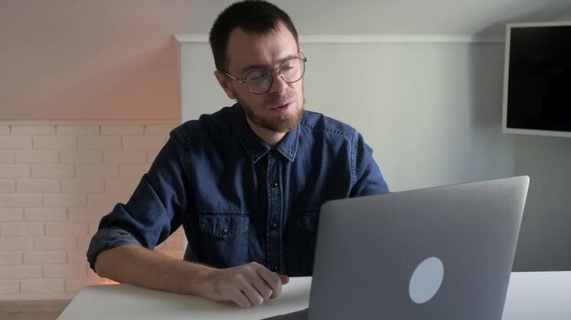 Young man in denim shirt giving thumbs-up and gesturing while speaking to laptop in modern home office, with man, male, adult, portrait detail, showing glass, beard, denim, shirt context