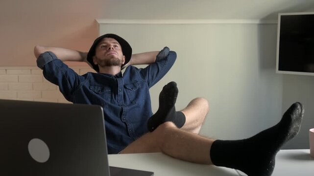 Young man seated at desk in modern minimal interior shifts through three visible states: initial relaxed pose with arms behind head, brief attentive lean toward laptop, then head-tilt back with eyes 