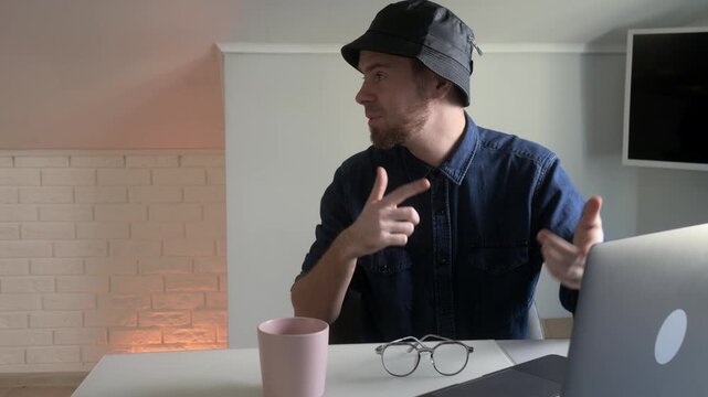 Young bearded man in hat interacting with laptop while seated at modern home office desk, with man, beard, hat, denim detail, showing shirt, laptop, typing, keyboard context