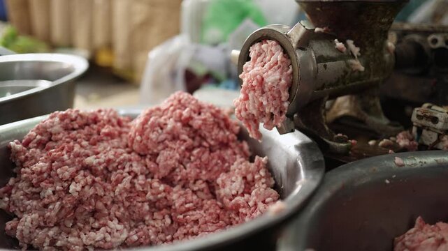 Close-up of a commercial meat grinder processing raw minced pork falling into a steel bowl at Xom Moi Market in Nha Trang, Vietnam. Food supply, butchery processing and retail market trade.