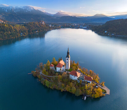 Aerial view of Bled Island and the Church of the Assumption rising above the calm waters of Lake Bled, surrounded by forested hills and the Julian Alps in Slovenia