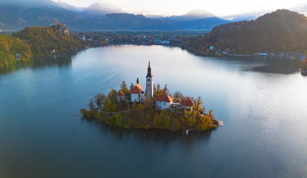 Aerial view of Bled Island and the Church of the Assumption rising above the calm waters of Lake Bled, surrounded by forested hills and the Julian Alps in Slovenia.