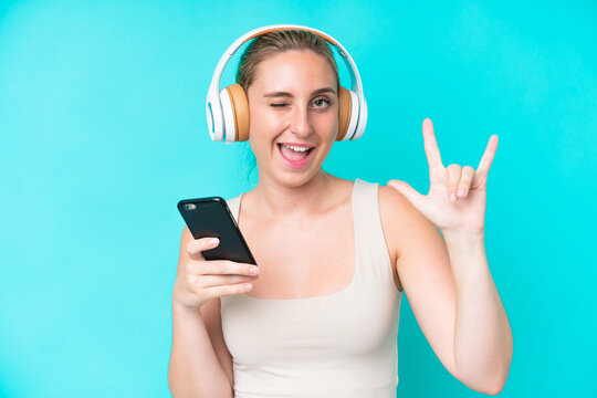 Young caucasian woman isolated on blue background listening music with a mobile making rock gesture