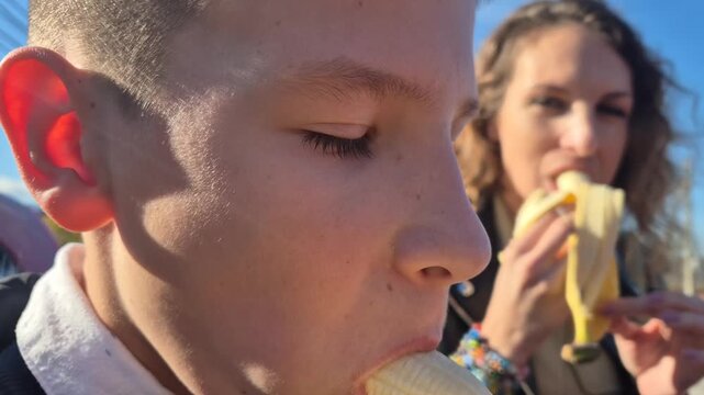 Young boy and mother eating fresh bananas outside on a sunny day. Handheld close up shot captures them taking bites.