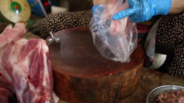 Close-up of a vendor in blue gloves shaving and preparing raw pork leg on a wooden chopping block at Xom Moi Market in Nha Trang, Vietnam. Commercial food supply and retail butchery trade.