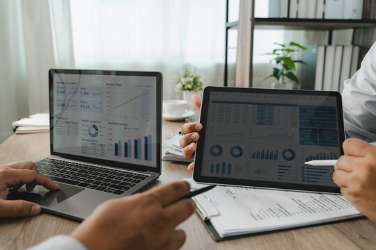 Business people reviewing financial performance data with a laptop dashboard, tablet, calculator and printed reports at an office desk, showing financial analysis and accounting workflow.