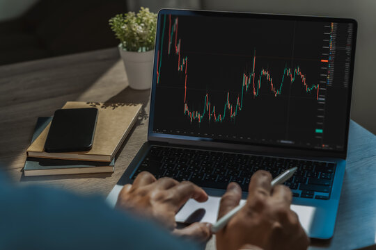 Businessman reviewing candlestick market chart on laptop holding a stylus, showing stock analysis, trading research and financial market monitoring at a desk.