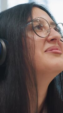 Woman with long dark hair wearing glasses and headphones. Dark locks frame subject against soft indoor backdrop. Setting appears calm with no distracting elements present