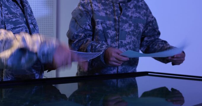 Military personnel in camouflage uniforms review documents and point at a digital map screen in a blue-lit command room.