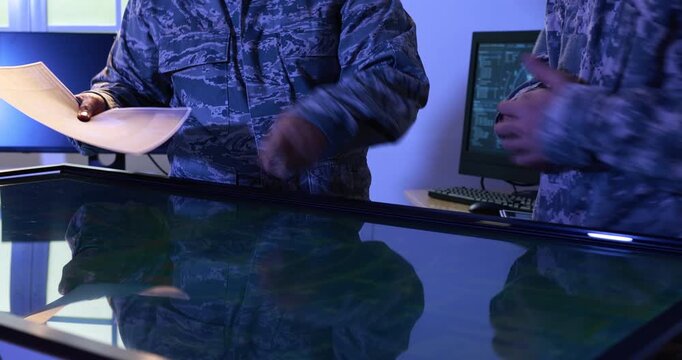 Military personnel in camouflage uniforms review documents and point at a large digital map table in a blue-lit command center.