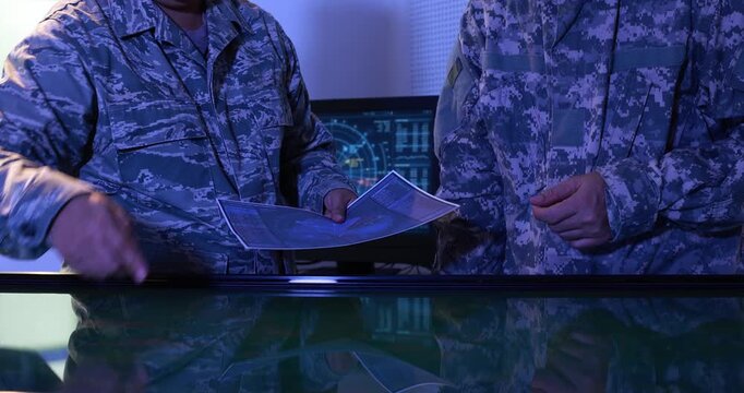 Two military personnel in camouflage uniforms review a paper map beside a glowing digital command table in a dark operations room.