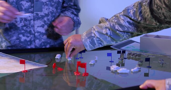 Military personnel in camouflage uniforms use a tabletop map with miniature flags and model vehicles during a strategy planning exercise.