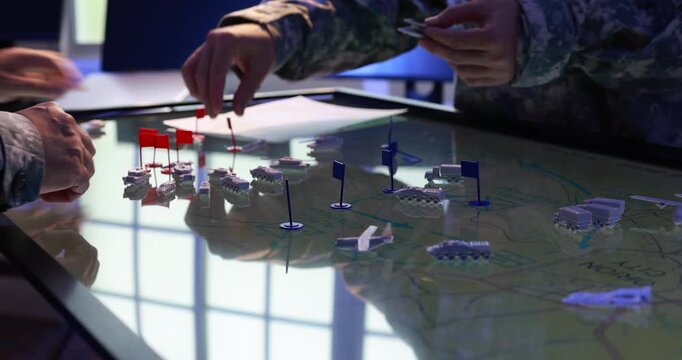 Military personnel in camouflage uniforms plan a tactical operation around an illuminated map table with miniature vehicles, ships and colored flags.