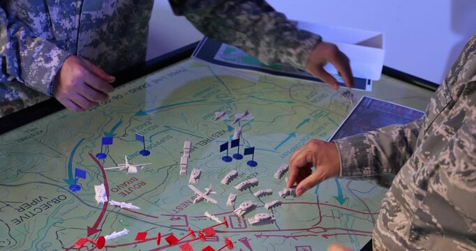 Military personnel plan a tactical operation on a large map using miniature vehicles, aircraft and colored markers in a command room.