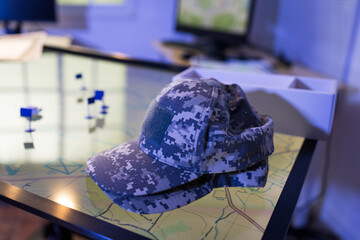 Naklejka premium Digital camouflage military cap resting on an interactive map display in a modern control room, with small flags and blurred monitors in the background.