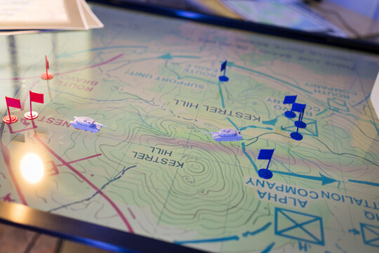 Close-up of a printed topographic map with red and blue pushpins marking routes, checkpoints and destinations under warm desk light.