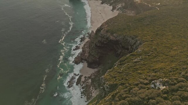 Drone view follows rugged coastal cliffs above turquoise ocean, foamy waves, secluded sandy coves and winding trails through vegetation, revealing expansive shoreline in warm evening light.