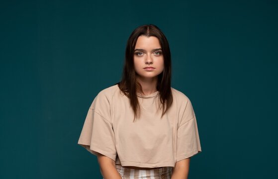 Young woman with neutral expression posing, wearing casual crop top against a teal background