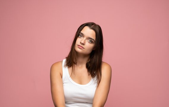 Young woman tilting head with an unamused expression, posing in a white tank top on a pink background