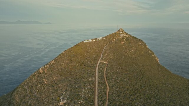 Drone aerial view of lighthouse atop green mountain ridge above calm sea, with narrow summit trail, rocky slopes, hazy horizon, and distant islands under soft daylight in serene setting.