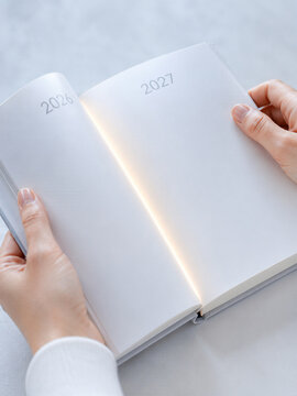 Woman hands holding open book with 2026 and 2027 numbers and glowing light between pages. Transition to new year 2027 concept. New year planning, fresh start and hope.